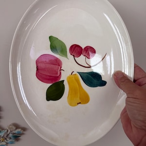 May include: An oval-shaped white ceramic serving platter featuring a hand-painted fruit motif in the center. The design includes a pink apple, a yellow pear, and two red cherries, all surrounded by several green leaves on a glossy off-white surface.
