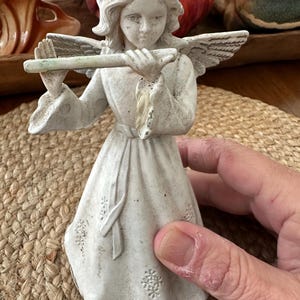 May include: White ceramic angel figurine playing a flute. The angel is wearing a long dress with a sash and has wings. The figurine is standing on a woven mat.