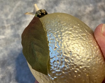 Op de afbeelding: Een goudkleurige, citroenvormige ornament met een getextureerd oppervlak. Het heeft een groen blad en een kleine stengel. Het ornament is waarschijnlijk voor feestdagen of seizoensgebonden decoratie.