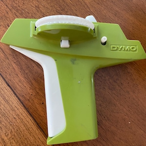 May include: A vintage green and white DYMO label maker. The label maker has a white handle and a green body with the word "DYMO" printed on the side. The label maker is made of plastic.