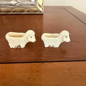 May include: Two small, cream-colored sheep-shaped planters with textured, curly wool detailing. Each figurine features a hollowed-out back for holding small items, with brown accents on the hooves and dark eyes on the sheep faces.