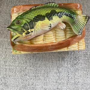 Könnte beinhalten: Ein dekoratives Keramikstück mit einem grün-schwarzen Bassfisch auf einem rechteckigen Sockel. Der Fisch ist mit Flossen und Schuppen detailliert, und der Sockel hat ein geflochtenes Korbmuster mit einem braunen Rand. Das Stück ist wahrscheinlich für die Inneneinrichtung.
