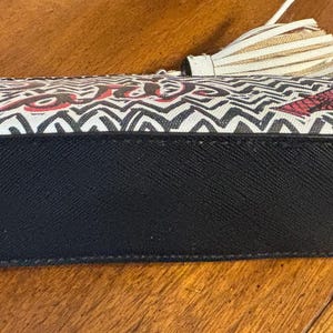 Puede incluir: Bolso de mano rectangular con base negra y estampado en zigzag blanco y negro. La palabra "love" est&aacute; escrita en rojo. El bolso tiene una borla.