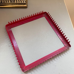 May include: A square red metal frame featuring a series of small, evenly spaced teeth or prongs along its outer edges. The frame is constructed from thin, flat strips of metal joined at the corners, creating a hollow center and a serrated perimeter.