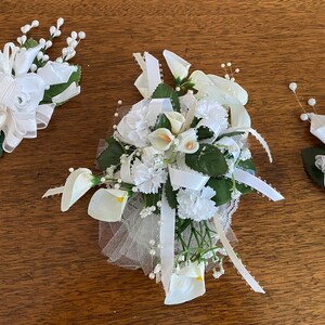May include: Three white floral arrangements on a wooden surface. The arrangements include calla lilies, roses, and other white flowers, with green leaves and white ribbons. One arrangement has a large bow.