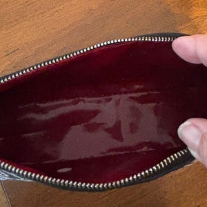 Puede incluir: Bolsa de cosm&eacute;ticos rectangular abierta, con cremallera negra e interior rojo. El exterior de la bolsa presenta un dise&ntilde;o estampado con detalles blancos, rojos y negros. El interior rojo brillante es visible.