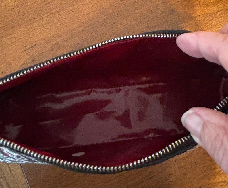 Puede incluir: Bolsa de cosm&eacute;ticos rectangular abierta, con cremallera negra e interior rojo. El exterior de la bolsa presenta un dise&ntilde;o estampado con detalles blancos, rojos y negros. El interior rojo brillante es visible.