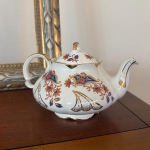 May include: A white porcelain teapot featuring a decorative pattern of blue and orange floral motifs, gold trim on the handle, spout, and lid, and a scalloped base. The teapot has a rounded body with a matching lid and a curved handle.