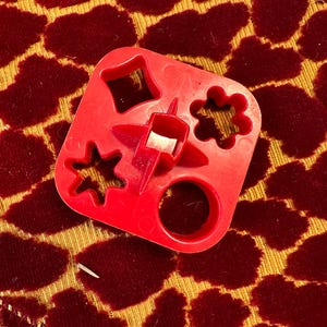 May include: Red plastic cookie cutter with four different shapes: a star, a bell, a circle, and a flower. The cutter is on a red and gold patterned surface.