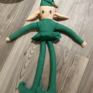 Puede incluir: Una muñeca de elfo de fieltro verde con un sombrero puntiagudo, orejas grandes y piernas largas. El elfo tiene una cara pintada con ojos azules y una sonrisa.