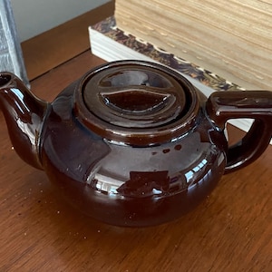 May include: A small, brown ceramic teapot with a matching lid. The teapot has a rounded body, a curved spout, and a handle. The glossy finish reflects light, and the teapot sits on a wooden surface.