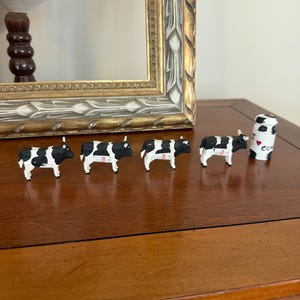 May include: Four small black and white cow figurines standing in a row, each with a red tag on its side, accompanied by a small white cylindrical container featuring a black and white cow print and the text I heart cows.