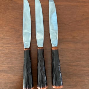 Puede incluir: Tres cuchillos de carne con hojas plateadas brillantes y mangos texturizados de color marrón oscuro. Los mangos tienen una apariencia de veta de madera. Los cuchillos están dispuestos verticalmente sobre una superficie marrón.