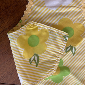May include: A close-up of a floral fabric with a yellow and white striped pattern. The fabric features large yellow flowers with green centers and leaves. A gold trim borders the edge of the fabric.