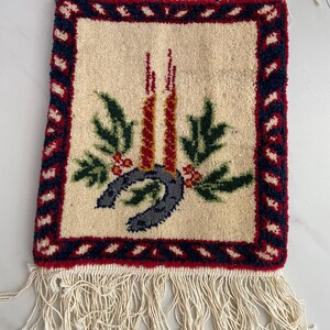May include: A rectangular, cream-colored rug with a festive design. The rug features a red and navy blue border, a central image of two red candles, green leaves, red berries, and a gray horseshoe. Cream-colored fringe hangs from the bottom.