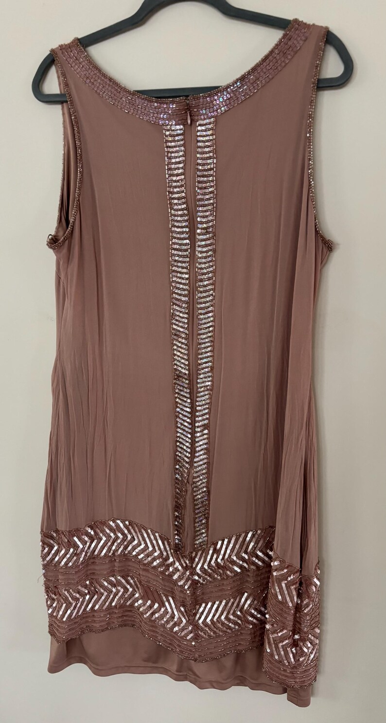 Peut inclure: Robe sans manches de couleur mauve avec des d&eacute;tails de sequins. La robe pr&eacute;sente un col orn&eacute; de sequins, une bande verticale de sequins au centre et un motif en chevrons &agrave; l'ourlet. Suspendue &agrave; un cintre noir.