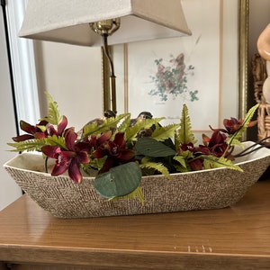 May include: A brown ceramic planter with a woven texture, filled with a floral arrangement of burgundy orchids and green ferns. The planter is sitting on a wooden surface.