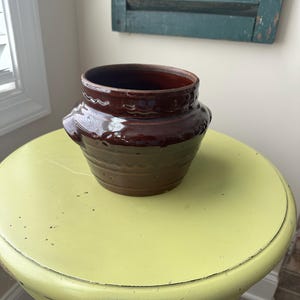 May include: A brown ceramic pot with a decorative design and two handles. The pot is sitting on a yellow table.
