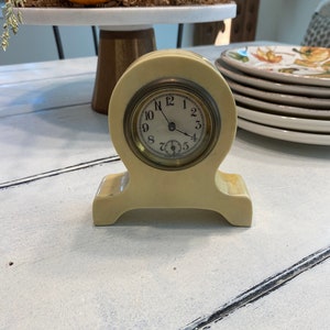 May include: A small, cream-coloured clock with a round face and black hands. The clock is on a white stand with a curved base. The clock face has black Roman numerals and a small black second hand.