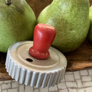 May include: A vintage metal cookie cutter with a red wooden handle, set against a backdrop of green pears. The cutter has a fluted edge and a small hole. The pears are a vibrant green color.