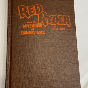 May include: A vintage hardcover book titled "Red Ryder and the Adventure at Chimney Rock." The book has a brown textured cover with orange lettering. The title is prominently displayed, with the author's name visible.