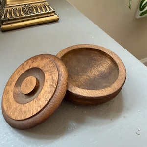 Può includere: Un contenitore rotondo in legno con coperchio abbinato, caratterizzato da una finitura con venature del legno marrone naturale. Il coperchio presenta un dettaglio circolare in rilievo al centro e la base è una semplice ciotola poco profonda.