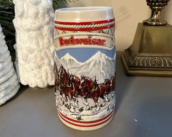 May include: A ceramic Budweiser beer stein with a white background and a red, white and blue design. The stein features a scene of a team of Clydesdale horses pulling a wagon through a snowy mountain landscape. The text "Budweiser" is printed in red on the stein.