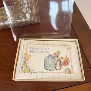May include: A rectangular photo album titled Grandma's Brag Book in a cream-colored box with a clear plastic lid. The cover features a classic illustration of a rabbit in a blue dress and a smaller rabbit, with floral accents and a small mouse.