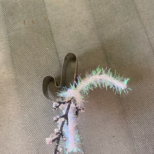 May include: A silver-colored metal cactus-shaped cookie cutter with a decorative sprig of white, iridescent, and black embellishments. The sprig has small, clear, square-shaped accents.