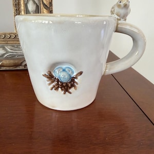 May include: A cream-colored ceramic mug featuring a small, sculpted bird perched on the handle. The front of the mug displays a raised, three-dimensional detail of a brown twig nest containing three speckled light blue eggs in a central indentation.