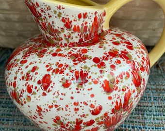May include: A ceramic pitcher with a white base, speckled with red and olive-green spots. It has a yellow handle and spout. The pitcher is likely used for serving beverages.