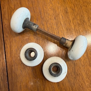 May include: A set of two white porcelain door knobs with a metal spindle connecting one pair, and two separate white porcelain circular rosettes with dark metal centres. The hardware features a smooth, glossy finish on the knobs and rosettes.