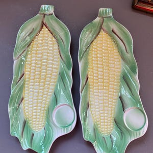 May include: Two ceramic corn-on-the-cob shaped serving dishes. The dishes are yellow and green, with a detailed design of corn kernels and leaves. Each dish has a small, round indentation. A charming addition to any kitchen.