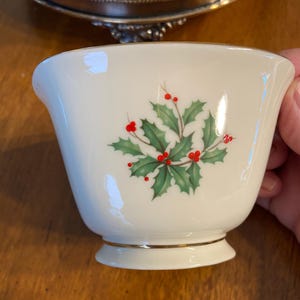 Könnte beinhalten: Cremefarbene Teetasse mit Goldrand und einem Stechpalmen- und Beeren-Design. Die Teetasse hat eine ausgestellte Form und einen kleinen Fuß. Die Stechpalmenblätter sind grün und die Beeren rot. Im Hintergrund befindet sich ein Silbertablett.