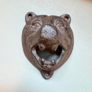Può includere: Un apribottiglie da parete in ghisa marrone a forma di testa di orso. L'orso presenta una superficie ruvida, orecchie visibili e una bocca aperta che funge da meccanismo di apertura. Sono presenti due fori per le viti di fissaggio.