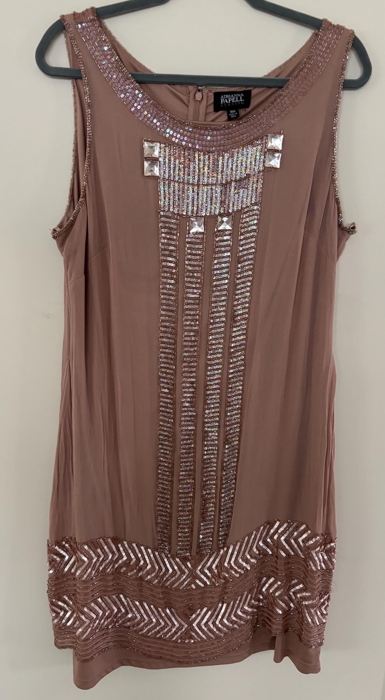 Peut inclure: Robe sans manches de couleur mauve orn&eacute;e de sequins et de perles. La robe pr&eacute;sente un d&eacute;collet&eacute; perl&eacute;, des motifs g&eacute;om&eacute;triques et des lignes verticales de sequins. La marque "Adrianna Papell" est visible sur l'&eacute;tiquette.
