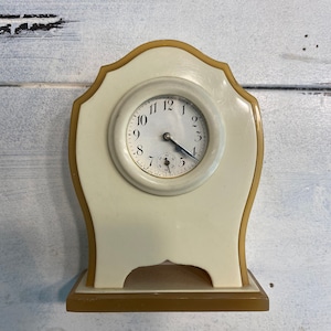 May include: A small, white, vintage clock with a gold trim and a round face. The clock has black Roman numeral hour markers and a black hand.
