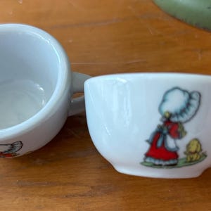 Peut inclure: Deux petites tasses à thé en céramique blanche. L'une est vide, l'autre présente une illustration de dessin animé d'une fille en robe rouge et d'un chien jaune. Les tasses ont de petites anses et sont posées sur une surface en bois.