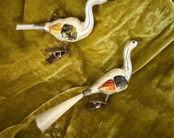 Può includere: Due ornamenti vintage a forma di uccello in vetro con colli lunghi, becchi neri e dettagli delle ali dipinti in arancione e nero. Ogni uccello ha una clip metallica fissata sul lato inferiore e una lunga coda in fibra chiara sul retro.