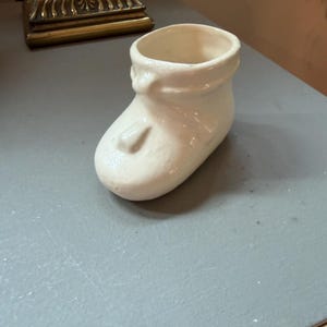 以下が含まれることがあります： オフホワイトの陶器製ベビーシューズ型容器。植木鉢や装飾品として使用可能。表面は滑らかで光沢があり、丸みを帯びた爪先、開口部にははっきりとした縁取り、そして甲の部分にはリボンやストラップを模した控えめな成形ディテールが施されている。