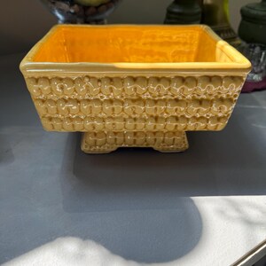 Könnte beinhalten: Eine quadratische gelbe Keramikschale mit einem erhabenen, sich wiederholenden Texturmuster an den Außenseiten. Die Schale hat eine glänzende Oberfläche, eine weite Öffnung und einen kleinen, integrierten Sockel, der den Hauptkörper anhebt.