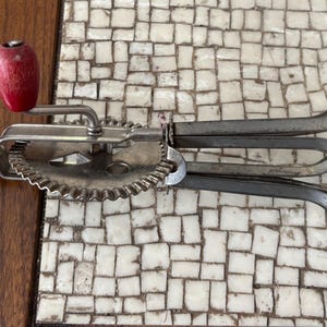 Può includere: Frusta per uova a manovella vintage con manici in legno rosso e crema. La frusta è in metallo e ha un meccanismo a ingranaggi. L'immagine mostra la frusta su una superficie di legno e una superficie di piastrelle a mosaico.