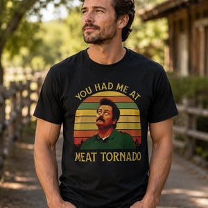 May include: A black short-sleeved t-shirt featuring a graphic print of a man with a mustache looking upward. The design includes a retro-style sunset background with horizontal stripes and the text "YOU HAD ME AT MEAT TORNADO" in yellow.