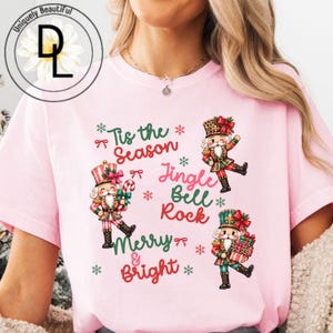 Può includere: Una t-shirt rosa chiaro con tre figure di schiaccianoci illustrate e il testo "Tis the Season", "Jingle Bell Rock" e "Merry & Bright". Il design include piccoli fiocchi rossi e fiocchi di neve verdi sparsi attorno agli schiaccianoci.