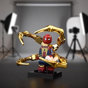Op de afbeelding: Een Spider-Man minifiguur in een rood en blauw pak met gouden accenten, staand op een zwarte basis. Het figuur is uitgerust met vier grote, metallic gouden mechanische armen die vanaf de rug uitsteken, elk voorzien van beweegbare gewrichten.
