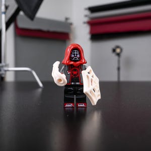 May include: A plastic minifigure featuring a red hood, a black and gray torso with a red spider emblem, and black legs. The figure wears a black mask with white eye details and holds a white plastic web accessory in one hand and a web shield in the other.