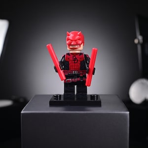 Peut inclure: Une figurine en plastique représentant un personnage portant un masque rouge à cornes et une combinaison rouge et noire avec un logo DD sur la poitrine. La figurine a une barbe et tient deux bâtons cylindriques rouges sur un socle noir.