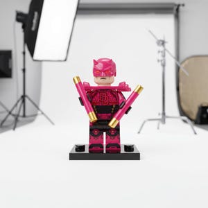 May include: A pink and dark red plastic minifigure featuring a horned mask, a chest emblem with the letters DD, and shoulder armor. The figure holds two pink batons with gold-colored tips and stands on a black rectangular display base.
