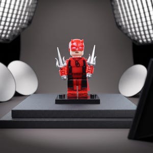 May include: A red and black plastic minifigure character wearing a horned mask and a suit with a DD emblem on the chest. The figure holds a silver-colored trident-style weapon in each hand and stands on a small black rectangular display base.