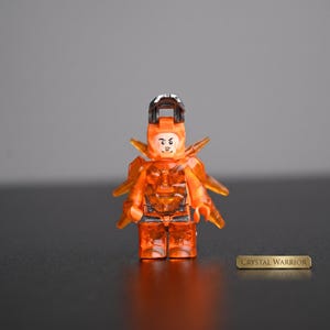 Puede incluir: Una minifigura de plástico naranja translúcido con un diseño detallado en el torso y las piernas, una pieza de armadura de hombro puntiaguda y un casco negro. La figura tiene una cara impresa con expresión seria y el texto CRYSTAL WARRIOR.